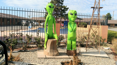 Two green alien statues stand on a stone base outside a fenced area, with a pool and buildings in the background.