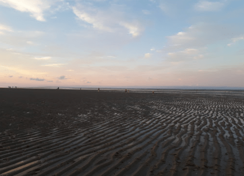 A serene beach at sunset, featuring rippled sand and a calm sea under a colorful sky with scattered clouds.