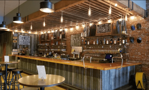 A rustic bar with wooden counters, shelves of bottles, and warm lighting, featuring metal stools and a cozy atmosphere.