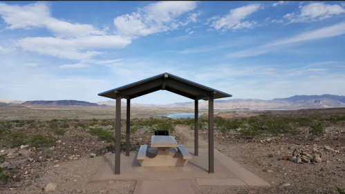 A shaded picnic area with a table overlooks a vast, arid landscape and distant mountains under a blue sky.