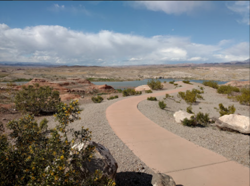 A winding path through a rocky landscape with sparse vegetation and a distant body of water under a cloudy sky.