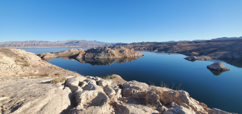 A serene landscape featuring calm blue water surrounded by rocky hills and mountains under a clear blue sky.