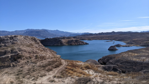 A panoramic view of a serene lake surrounded by rocky hills and distant mountains under a clear blue sky.
