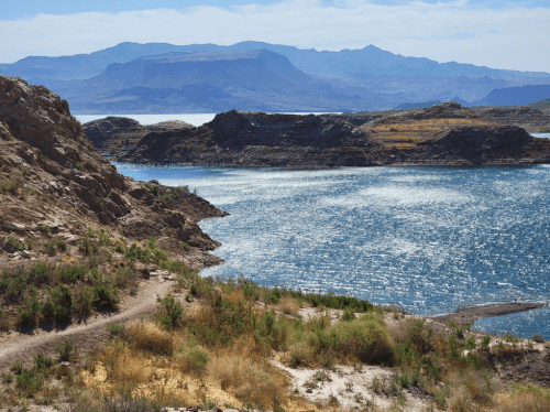 A serene landscape featuring a shimmering lake surrounded by rocky hills and distant mountains under a clear blue sky.