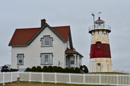 A weathered lighthouse beside a historic house, both surrounded by a white picket fence under a cloudy sky.