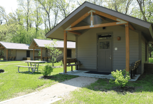 A cozy cabin with a porch, surrounded by greenery, and picnic tables in a serene outdoor setting.