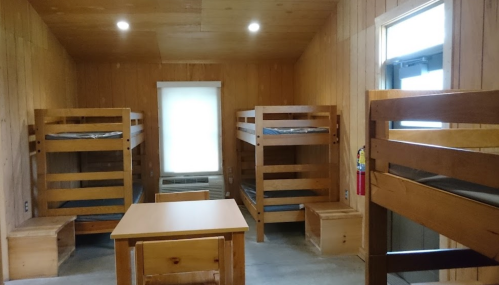 A simple cabin interior with two sets of wooden bunk beds, a table, and a window, all in a wooden-paneled room.