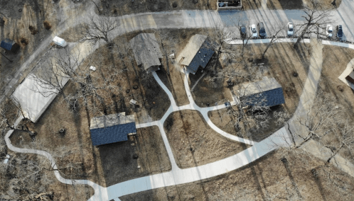 Aerial view of a park with winding paths, several buildings, and parked cars, surrounded by bare trees.