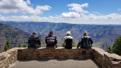 Four people sit on a stone wall overlooking a vast canyon landscape under a blue sky with clouds.