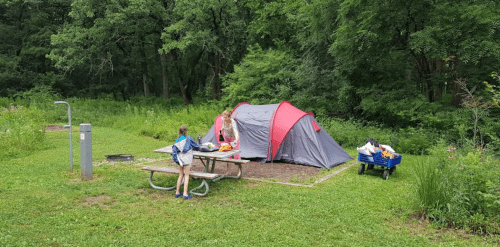 A campsite with a red and gray tent, a picnic table, and a child near a wagon filled with supplies, surrounded by greenery.