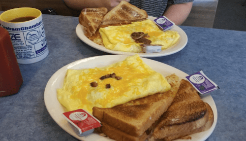 Two plates of breakfast featuring omelets, toast, and syrup packets on a blue table.