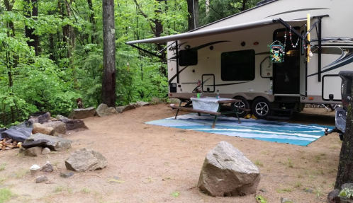 A camper parked in a wooded area, with a picnic table and a rug on the ground surrounded by rocks and greenery.