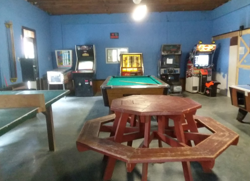 A game room featuring a pool table, arcade machines, and a large wooden picnic table in a blue-walled space.