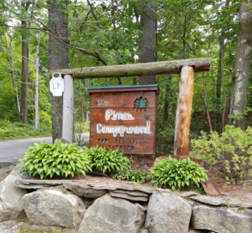 Wooden sign for "Pine Campground" surrounded by trees and greenery, with a stone base and a road in the background.
