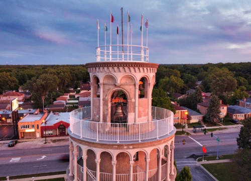 Aerial view of a tall tower with flags on top, surrounded by trees and buildings under a colorful sky.