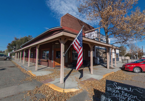 A corner building with a porch, featuring an American flag and autumn leaves scattered on the ground.