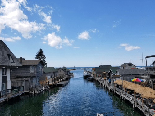A scenic view of a calm waterway lined with wooden buildings, leading to a marina under a clear blue sky.
