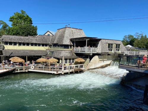 A riverside building with a deck, umbrellas, and flowing water, under a clear blue sky.