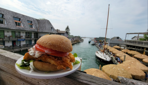 A fried fish sandwich with tomato and mayo on a bun, set against a scenic waterfront with boats and thatched umbrellas.