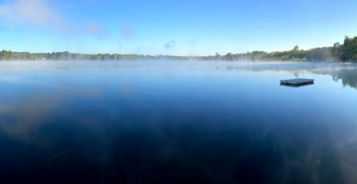 A serene lake at dawn, with mist rising from the water and a small dock floating in the distance.