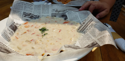 A bowl of creamy seafood chowder served in a dish lined with newspaper, with a hand reaching for it.
