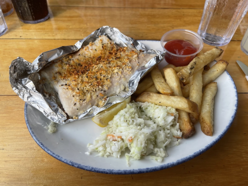 A plate with baked fish in foil, coleslaw, fries, and a small cup of ketchup on a wooden table.