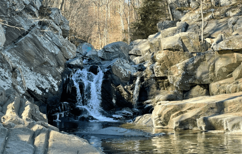 A serene waterfall cascades over rocky terrain, surrounded by trees and reflecting in a calm pool of water.