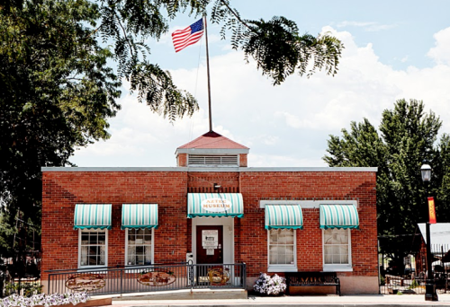 A brick building with green awnings, featuring a flagpole and sign for a museum, surrounded by trees and flowers.