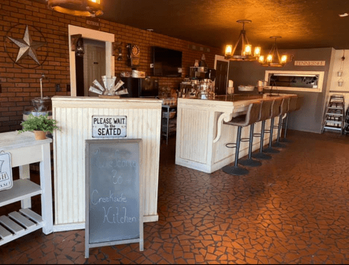 Cozy restaurant interior with a wooden bar, seating, and a welcome sign. Warm lighting and rustic decor create a welcoming atmosphere.