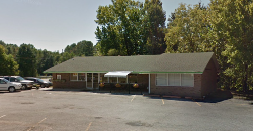 A single-story brick building surrounded by trees, with a parking lot in front and a covered entrance.