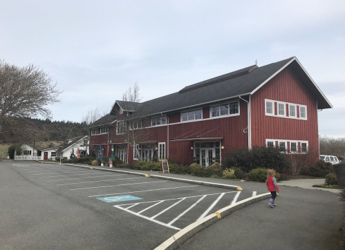 A red barn-style building with large windows, surrounded by greenery and a parking lot, on a cloudy day.