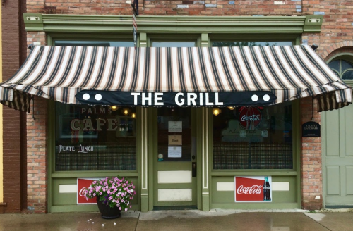 A storefront with a striped awning reading "THE GRILL," featuring flower pots and Coca-Cola signage.