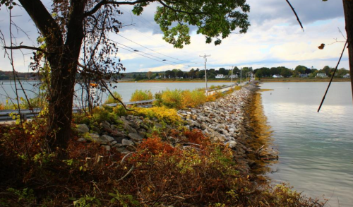 A rocky path extends into a calm body of water, framed by trees and autumn foliage under a cloudy sky.