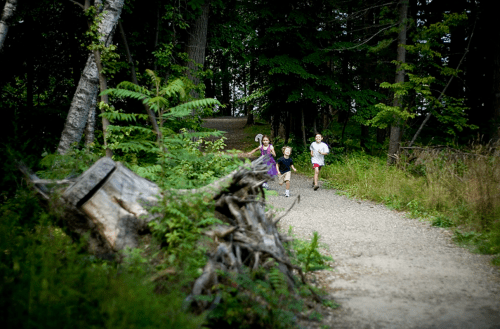 Three children running along a gravel path in a wooded area, surrounded by trees and greenery.