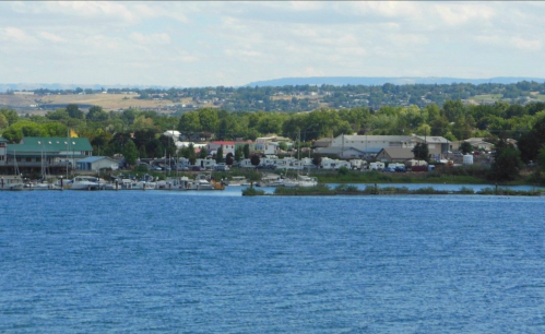A scenic view of a lake with boats, surrounded by greenery and buildings in the background under a blue sky.