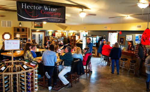 A bustling wine tasting room with people socializing, a bar, and shelves of wine bottles in a cozy setting.