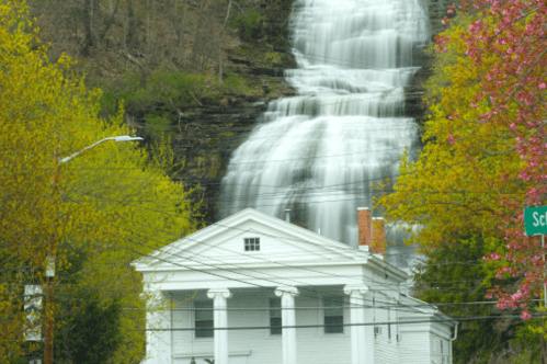 A white house with columns in front of a cascading waterfall, surrounded by vibrant green trees and pink blossoms.