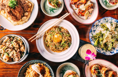 A vibrant spread of various dishes, including noodles, fried chicken, salads, and appetizers, arranged on a wooden table.