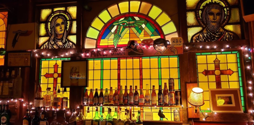 Stained glass windows with religious figures above a bar shelf lined with various bottles and warm lighting.