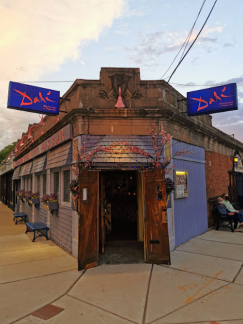 Exterior of a restaurant with a blue facade, wooden doors, and colorful signage, surrounded by flower pots and street benches.