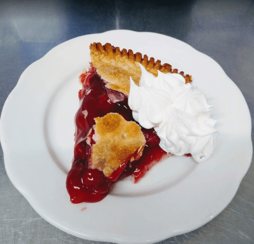 A slice of cherry pie with a flaky crust, topped with a dollop of whipped cream on a white plate.