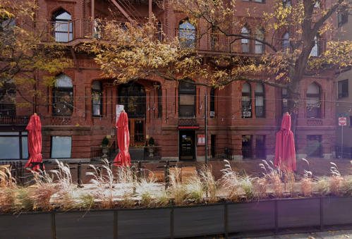 A brick building with large windows, surrounded by trees and outdoor seating with red umbrellas.