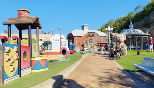 Colorful playground with themed structures, a large teddy bear, and families enjoying a sunny day.