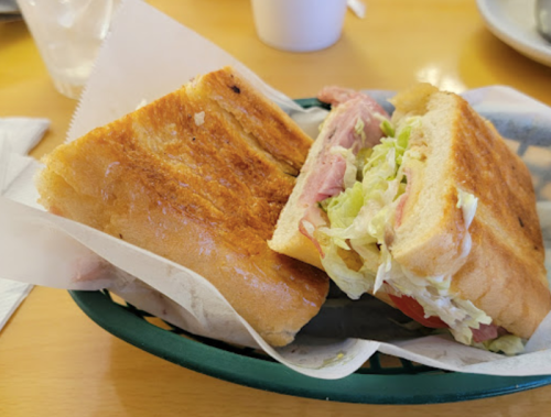 A toasted sandwich with ham, lettuce, and tomato, served in a green basket on a wooden table.