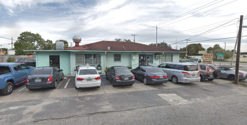 A small, light blue building with a red roof, surrounded by parked cars, under a cloudy sky.