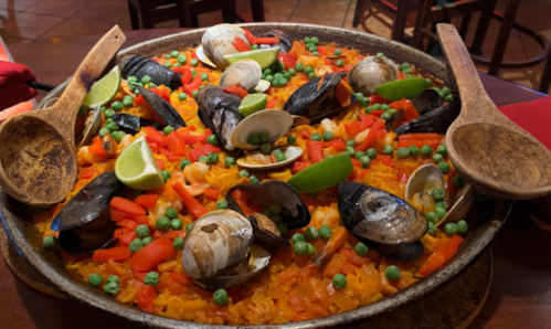 A large paella dish filled with seafood, vegetables, and garnished with lime wedges, served with wooden spoons.