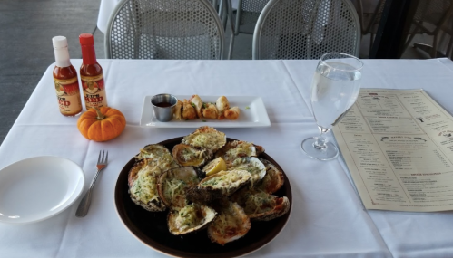 A plate of baked oysters with lemon, a small dish of sauce, hot sauce bottles, a glass of water, and a menu on a table.