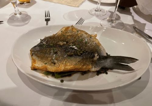 A whole cooked fish garnished with capers, served on a white plate with a side of greens.
