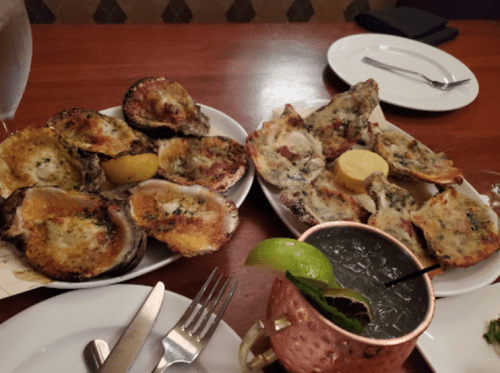 Two plates of baked oysters with lemon, alongside a copper mug cocktail, on a wooden table.