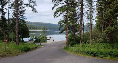 A serene lake view surrounded by trees, with a boat dock and a person in the distance on a calm day.